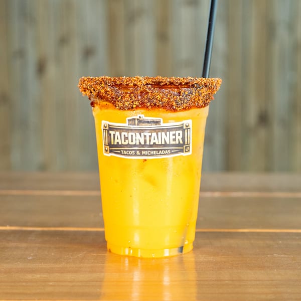 A Tacontainer Michelada, a yellow drink with a chili-lime rim, sits on a wooden table.