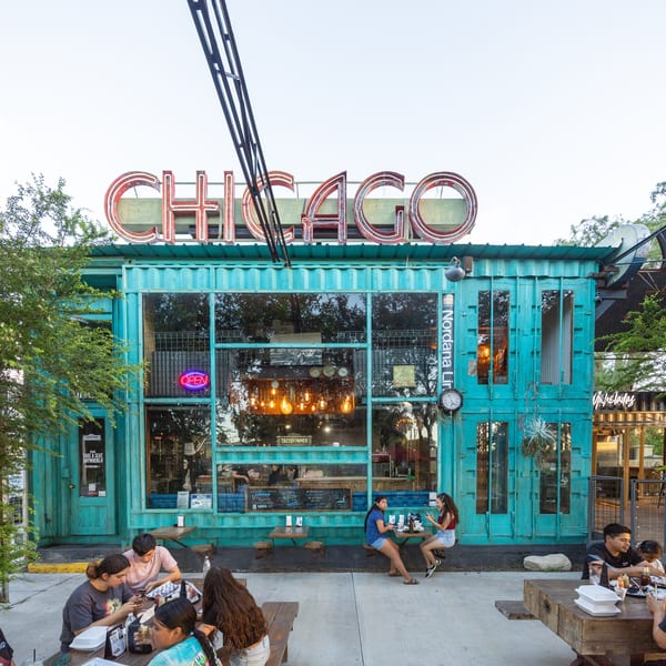 People dining outdoors at picnic tables in front of a turquoise restaurant with a large "CHICAGO" sign.
