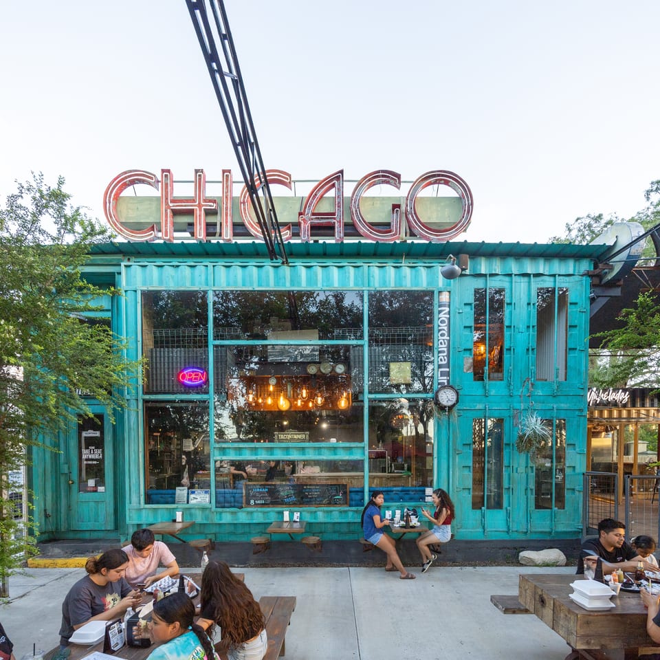 People dining outdoors at picnic tables in front of a turquoise restaurant with a large "CHICAGO" sign.