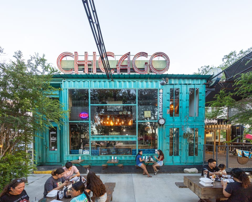 People dining outdoors at picnic tables in front of a turquoise restaurant with a large "CHICAGO" sign.