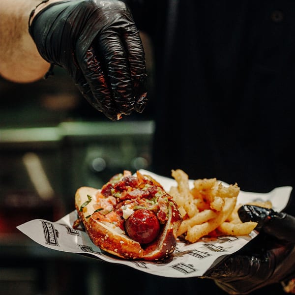 A chef in black gloves sprinkles seasoning on a loaded hot dog with fries.