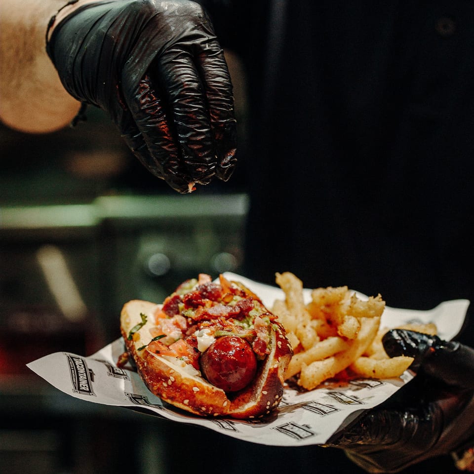 A chef in black gloves sprinkles seasoning on a loaded hot dog with fries.