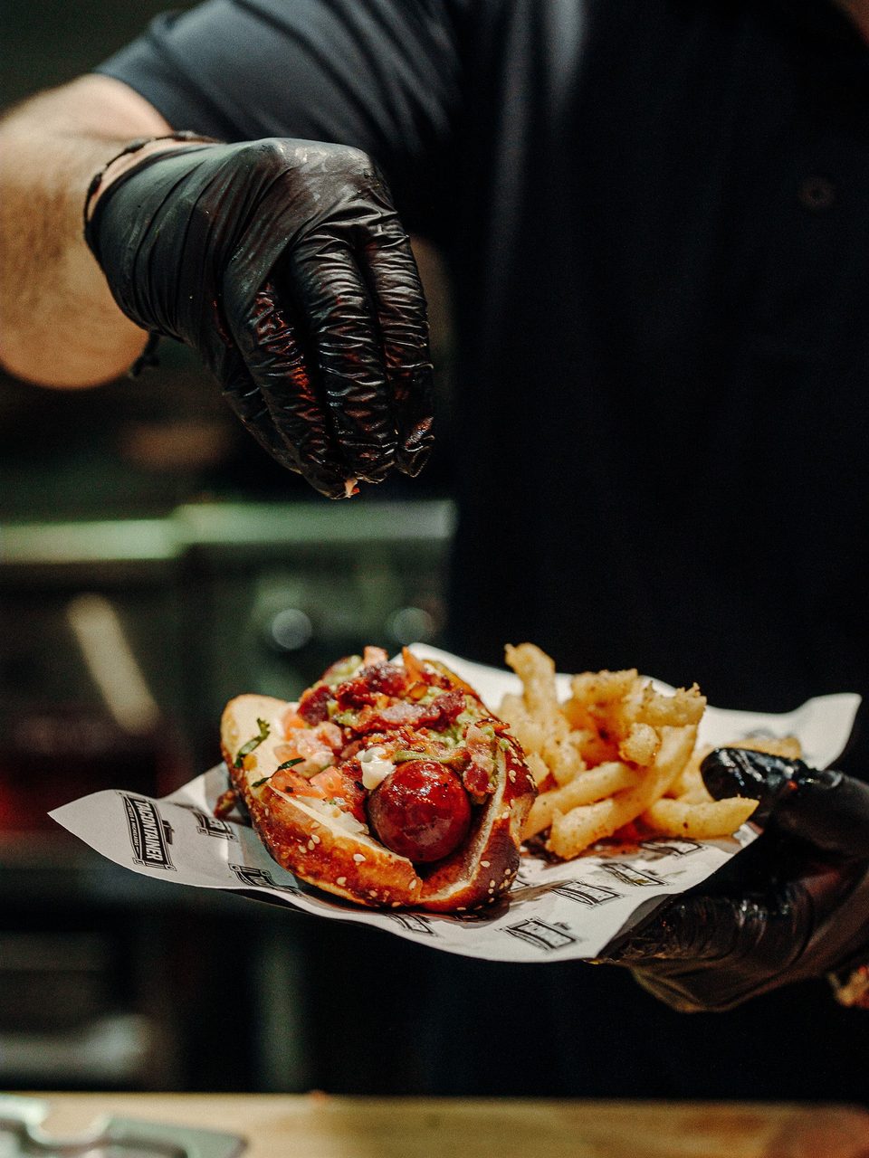 A chef in black gloves sprinkles seasoning on a loaded hot dog with fries.