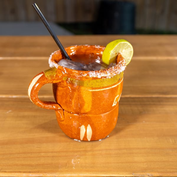 A traditional Mexican cantarito cocktail with a salted rim and lime wedge, on a wooden table.