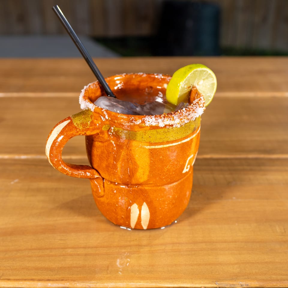 A traditional Mexican cantarito cocktail with a salted rim and lime wedge, on a wooden table.