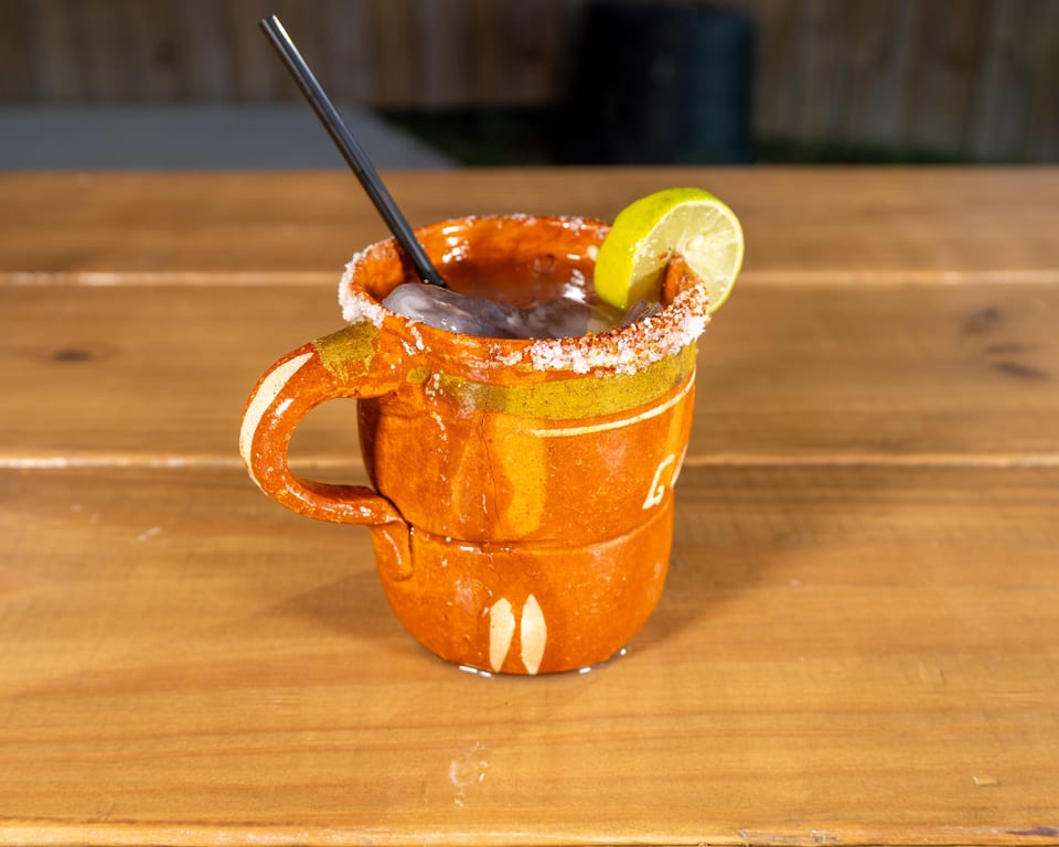 A traditional Mexican cantarito cocktail with a salted rim and lime wedge, on a wooden table.