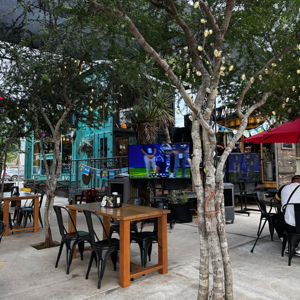 Outdoor patio with wooden tables, black chairs, string lights on trees, and TVs showing sports.
