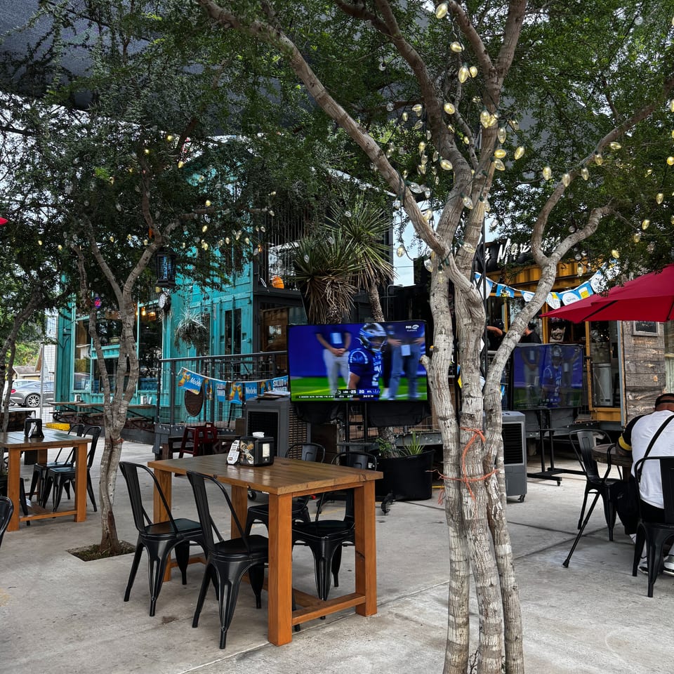 Outdoor patio with wooden tables, black chairs, string lights on trees, and TVs showing sports.