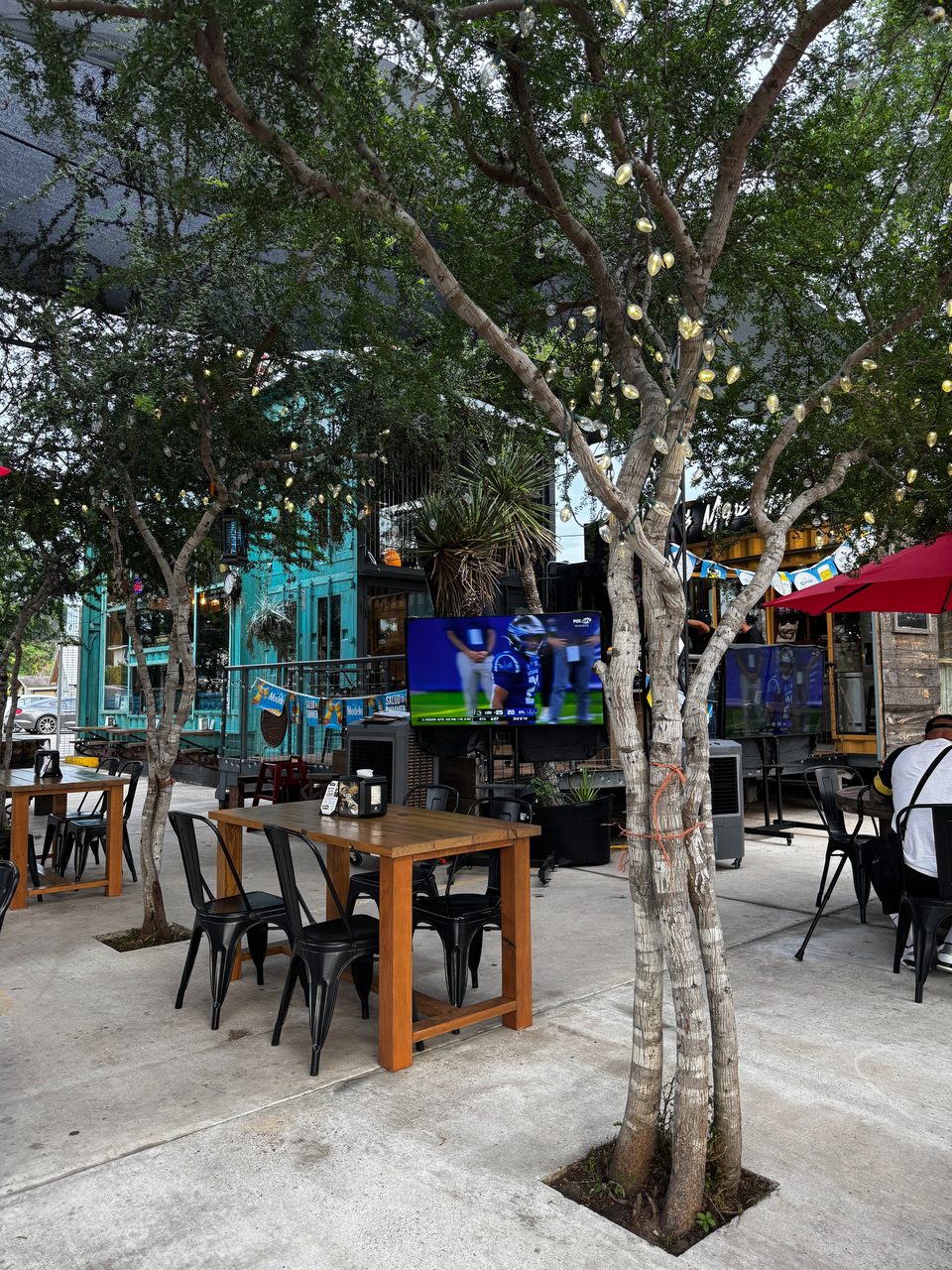Outdoor patio with wooden tables, black chairs, string lights on trees, and TVs showing sports.