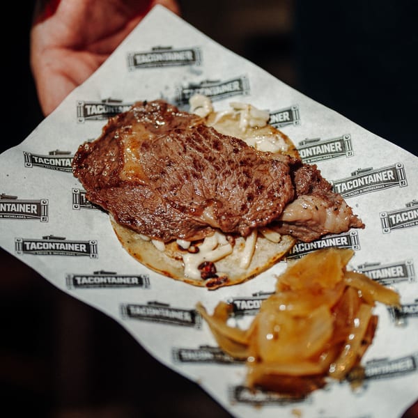 Hand holding a TaconTainer taco with grilled steak, white sauce, and caramelized onions.