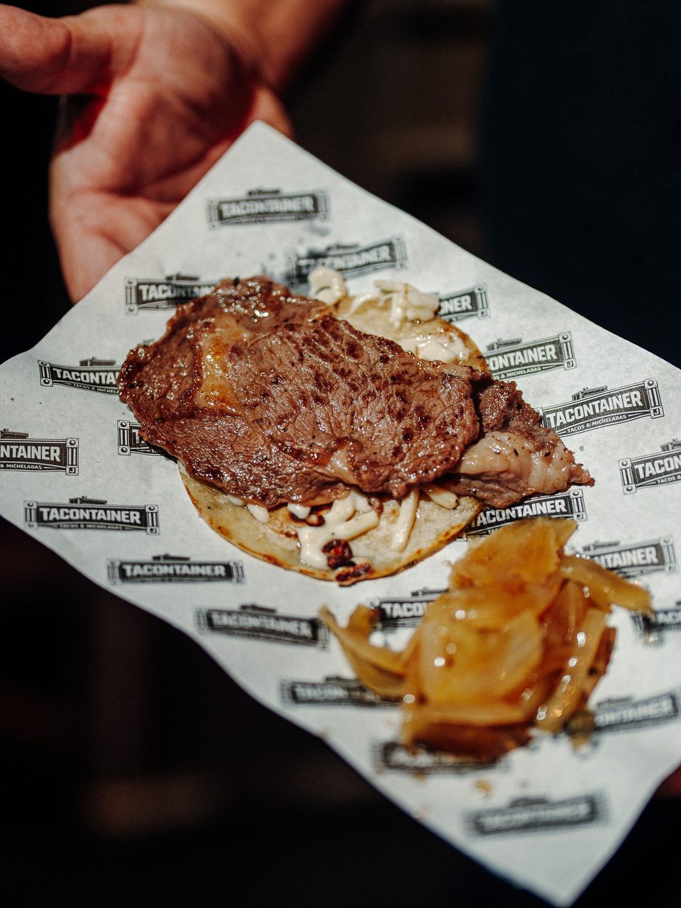 Hand holding a TaconTainer taco with grilled steak, white sauce, and caramelized onions.