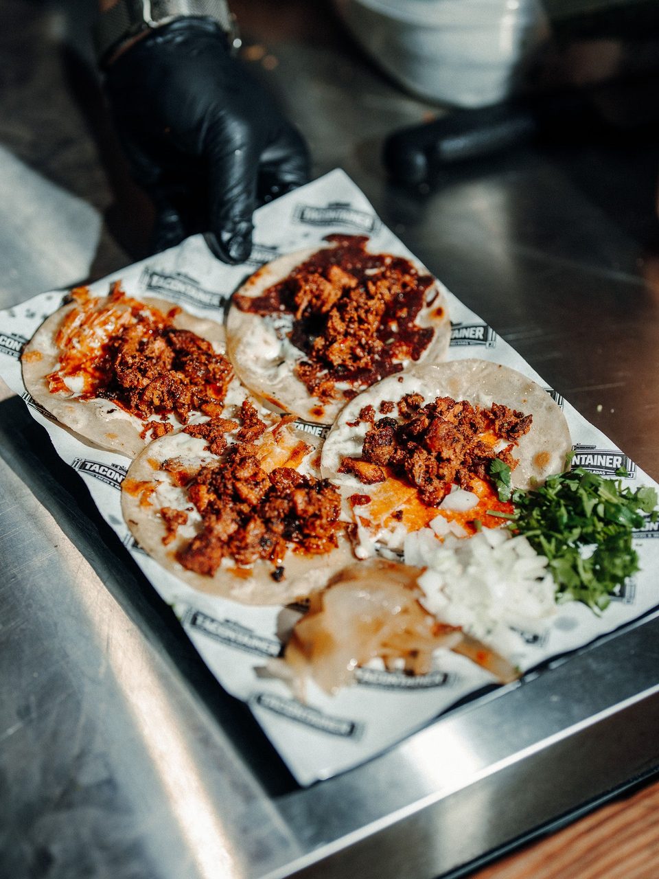 A gloved hand holds a tray of four tacos with meat, sauce, cilantro, and onions on a metal surface.