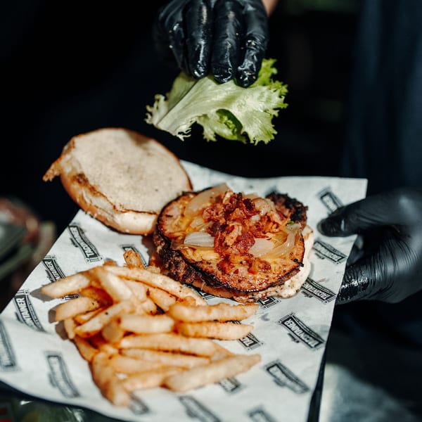 Gloved hands assemble a burger with bacon, onions, and lettuce, served with fries on branded paper.