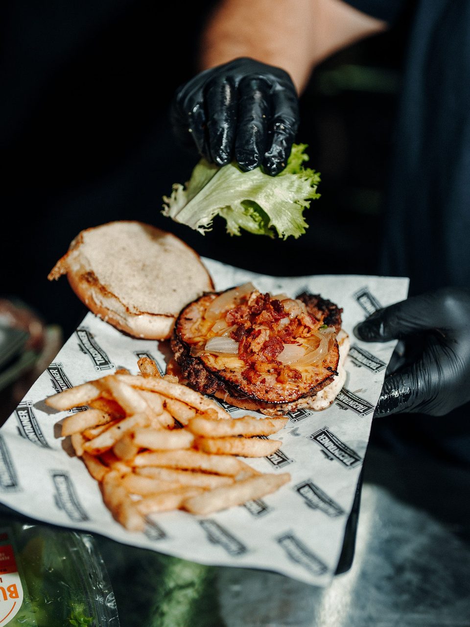 Gloved hands assemble a burger with bacon, onions, and lettuce, served with fries on branded paper.