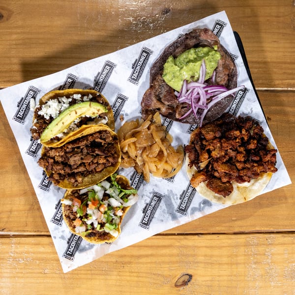Assortment of TACONTAINER tacos with various toppings, including avocado, cheese, and onions, on a wooden table.