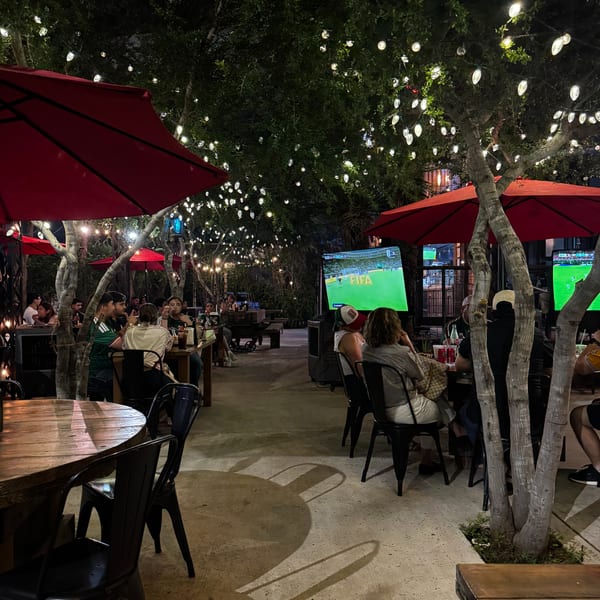 Outdoor restaurant patio at night with patrons watching soccer on large screens under string lights.