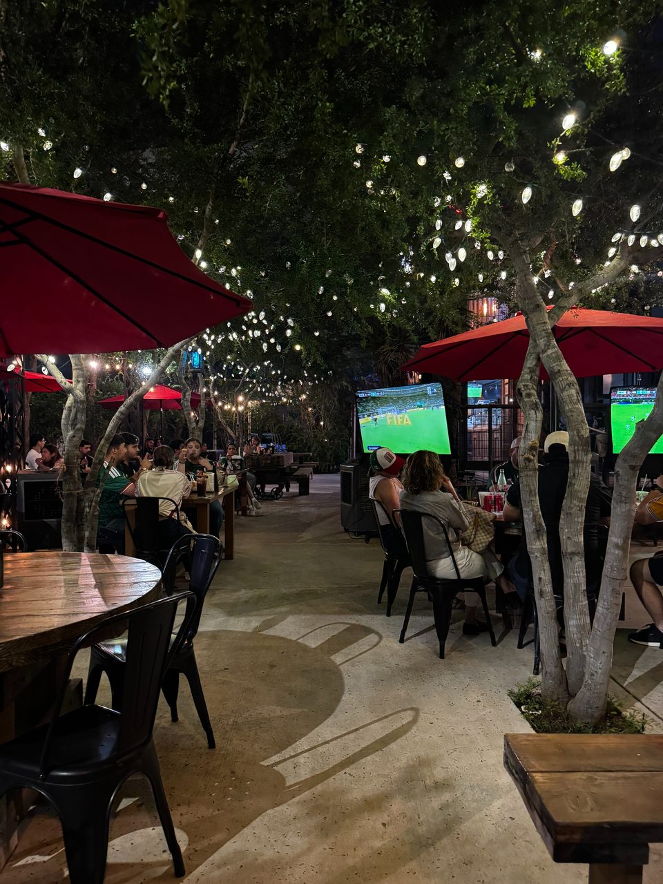Outdoor restaurant patio at night with patrons watching soccer on large screens under string lights.