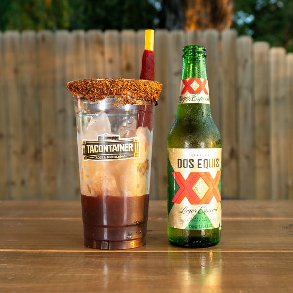 A Tacontainer Michelada with a tamarind candy straw next to a cold bottle of Dos Equis beer.