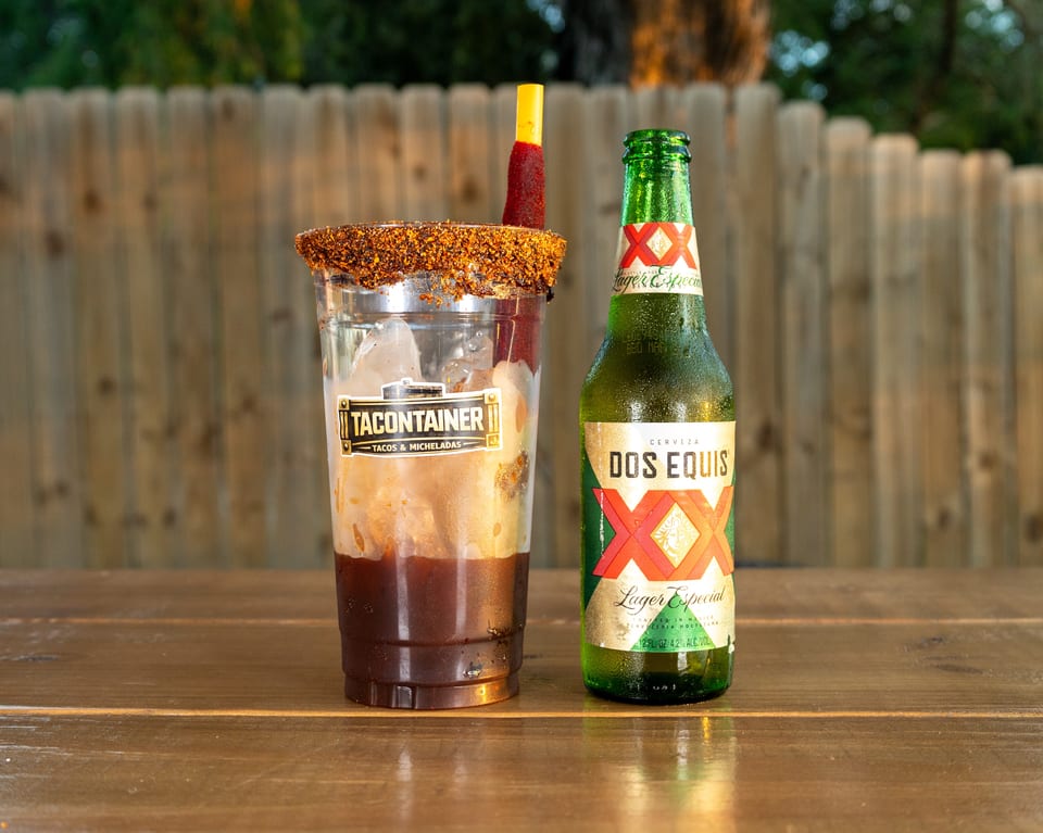 A Tacontainer Michelada with a tamarind candy straw next to a cold bottle of Dos Equis beer.