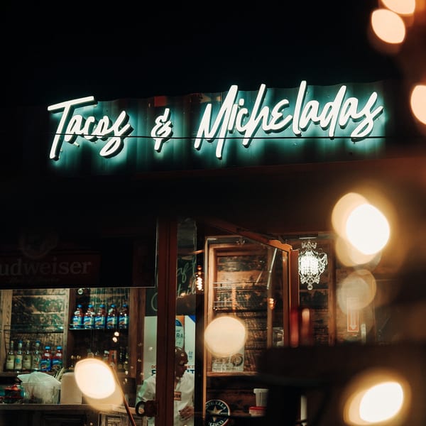 Neon sign for "Tacos & Micheladas" glowing above a restaurant entrance at night, with bokeh lights.