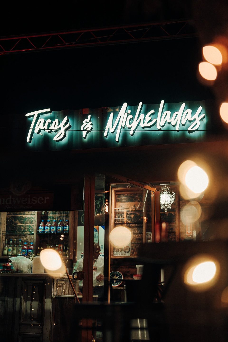 Neon sign for "Tacos & Micheladas" glowing above a restaurant entrance at night, with bokeh lights.